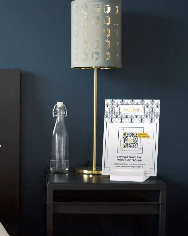 Hotel bedside table with a bottle of water, lamp, and sign asking for reviews on it.