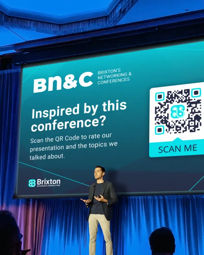 A man presenting at a conference in front of a projector screen. The screen shows a QR Code that allows scanners to rate the presentation.