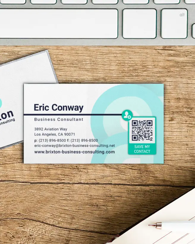 The front of a business card and the back of a business consultant's business card with contact information and a QR Code on it lying on a wooden desk.
